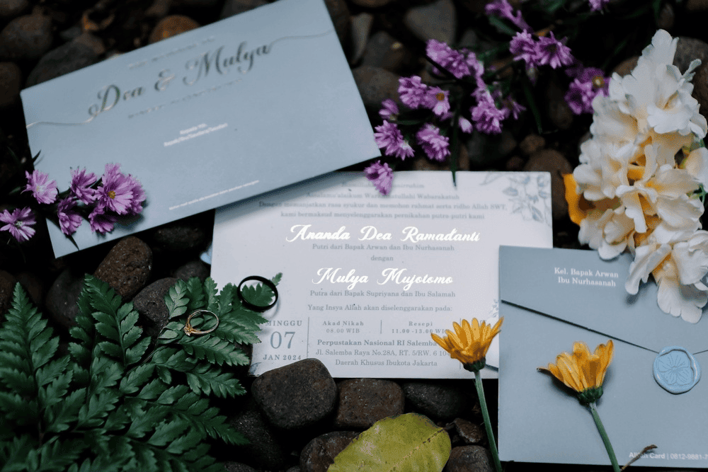 • A dusty blue wedding invitation suite with gold script lettering, surrounded by purple and white flowers on dark stones.