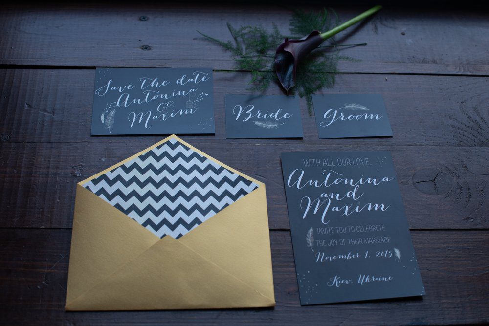 Charcoal grey wedding invitation suite with white calligraphy, a gold envelope, and a feather illustration on a dark wooden table.
