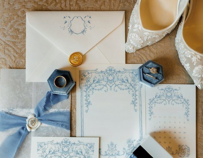Vintage-style wedding invitation suite with pale blue ornamental borders, a gold wax seal, blue velvet ring boxes, and lace bridal shoes.