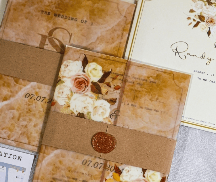 A rustic kraft paper wedding invitation suite featuring a marbled texture background, cream-colored floral illustrations, a brown paper belly band, and a round bronze wax seal.